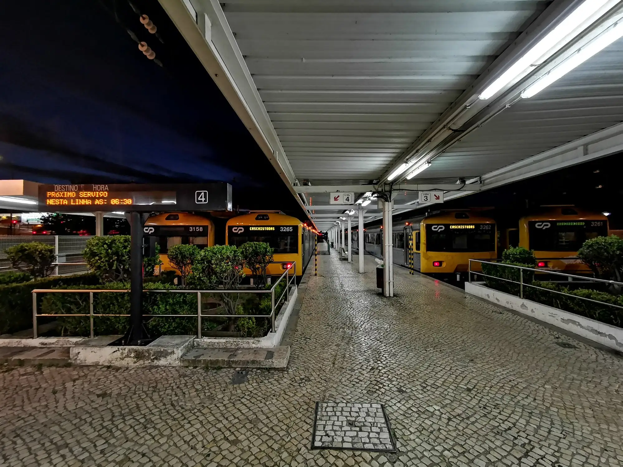 An empty platform at Cascais railway station at night, with a departure board and yellow suburban trains