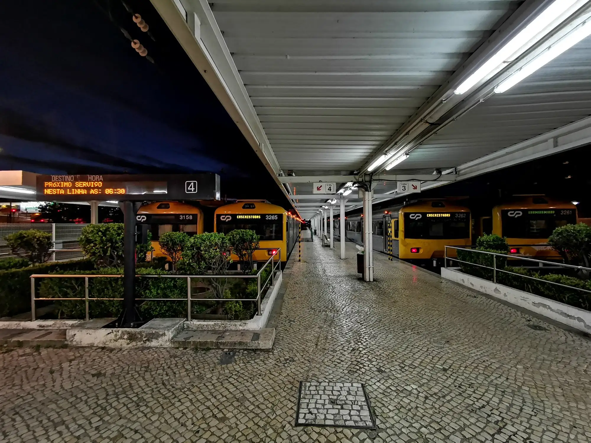 An empty platform at Cascais railway station at night, with a departure board and yellow suburban trains